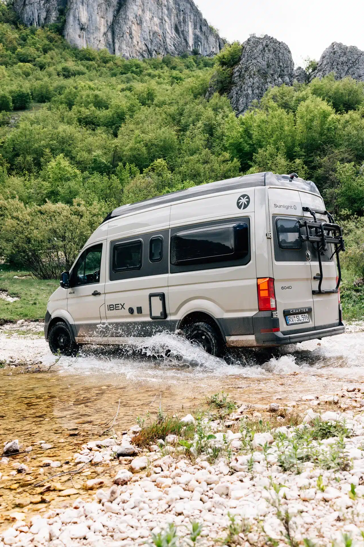 Sunlight IBEX VW 604D Allrad-Camper beim Durchqueren eines flachen Bachs vor felsiger und bewaldeter Kulisse.