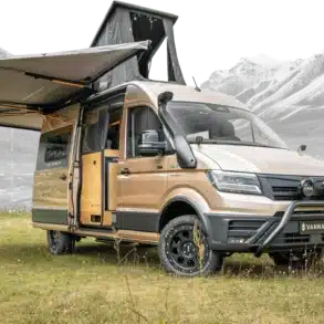 Vannado Karakorum Expeditionscamper, beige, mit Markise und Aufstelldach, steht auf einer Wiese vor Bergen.