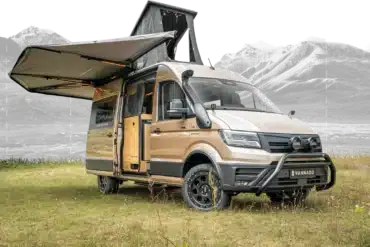 Vannado Karakorum Expeditionscamper, beige, mit Markise und Aufstelldach, steht auf einer Wiese vor Bergen.