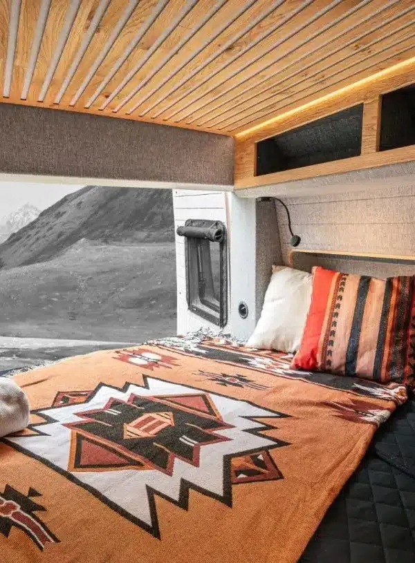 Schlafbereich im Vannado Karakorum Campervan mit Naturholz-Verkleidung, festem Bett und Kissen, Blick zum Fenster und Berge draußen