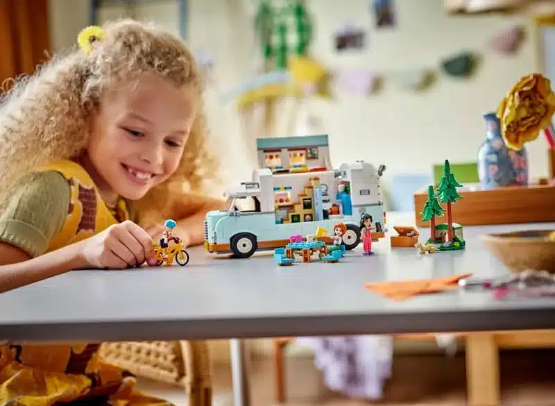 Kind spielt mit dem LEGO Friends Wohnmobil 42663, Spielfiguren, Fahrrad und Campingzubehör auf einem Tisch.