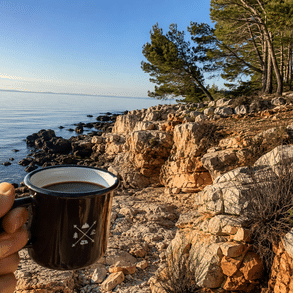 Hand hält eine Tasse Kaffee vor einer felsigen Küste und dem ruhigen Meer.