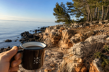 Hand hält eine Tasse Kaffee vor einer felsigen Küste und dem ruhigen Meer.