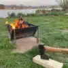 Schwarzer Grill und Feuerschale aus Stahl mit brennendem Holzfeuer auf einer Wiese am See