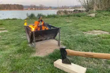 Schwarzer Grill und Feuerschale aus Stahl mit brennendem Holzfeuer auf einer Wiese am See