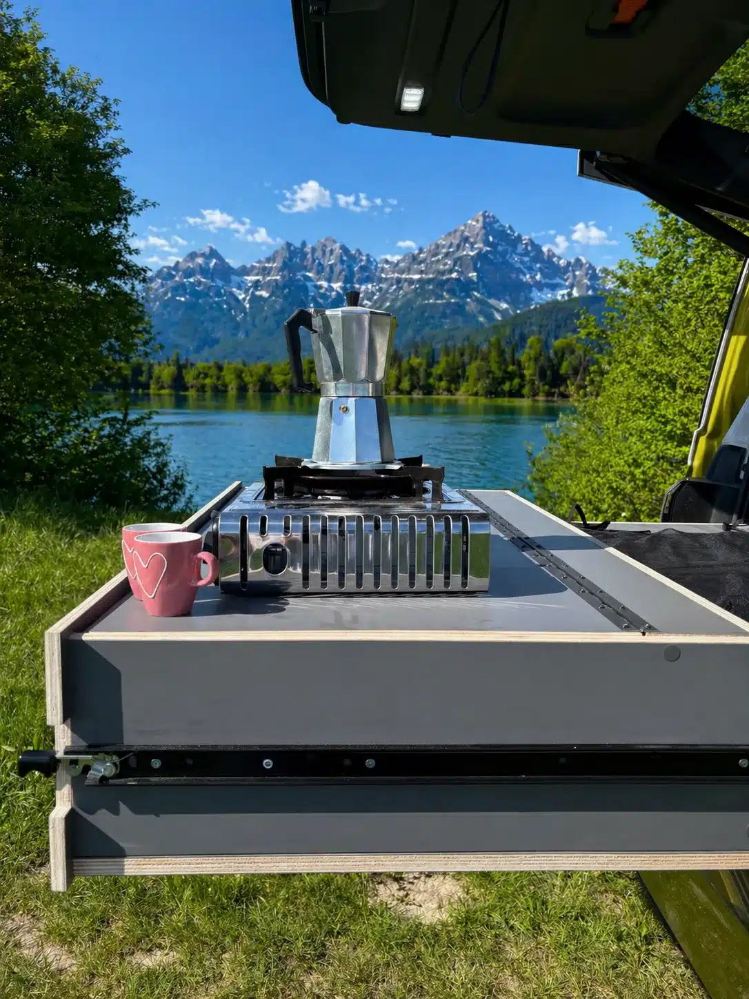 Campingbox S KISTL Star mit Herd, Kaffeekanne und Tasse an einem See mit Bergkulisse.