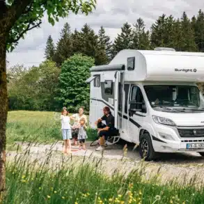 Familie steht und sitzt vor einem Sunlight Alkoven Root A60 Wohnmobil auf einer Wiese