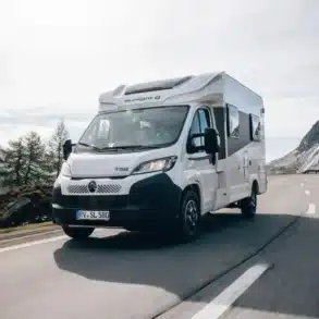 Weißer Sunlight T 58 Root fährt auf einer kurvenreichen Bergstraße vor verschneiten Alpen