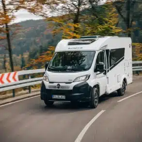 Weißer Sunlight V 60 Root fährt auf einer kurvenreichen Straße durch einen herbstlichen Wald.