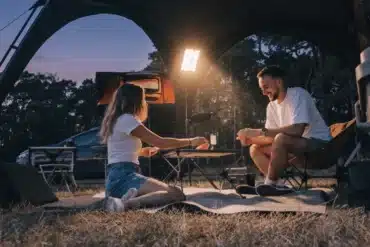 Campingpaar sitzt abends neben einem Fahrzeug mit Dachzelt an Campingtisch und Campingstühlen unter einer Stativleuchte