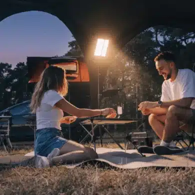 Campingpaar sitzt abends neben einem Fahrzeug mit Dachzelt an Campingtisch und Campingstühlen unter einer Stativleuchte