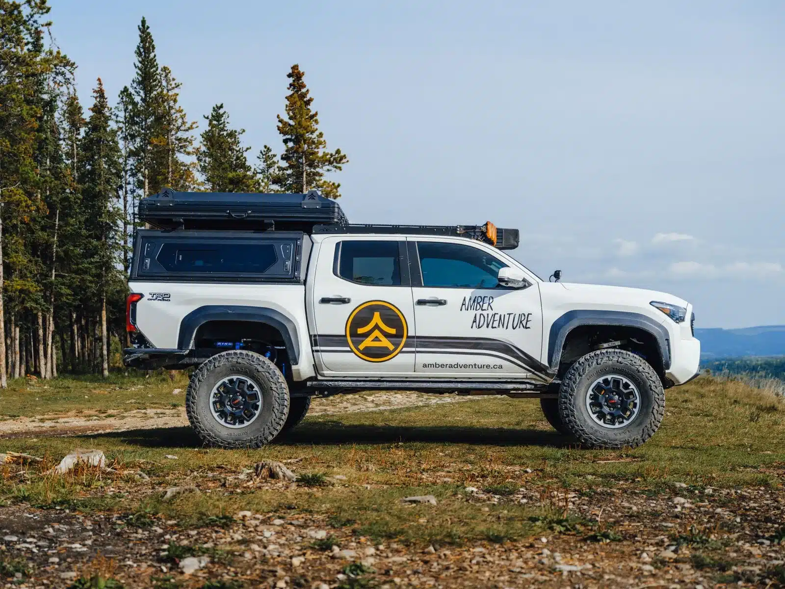Pickup mit schwarzem Aluminium-Hardtop und The Cabin 2.0 XL Dachzelt, geparkt auf einer Lichtung vor Bäumen