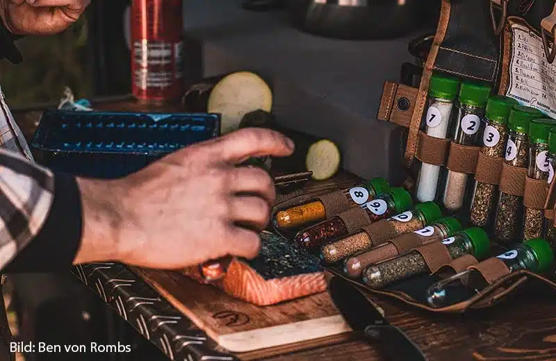 Eine ausgerollte Tool Roll Spice mit nummerierten Gewürzbehältern und einer Hand, die Gewürze zum Würzen von frischem Lachs beim Camping verwendet.