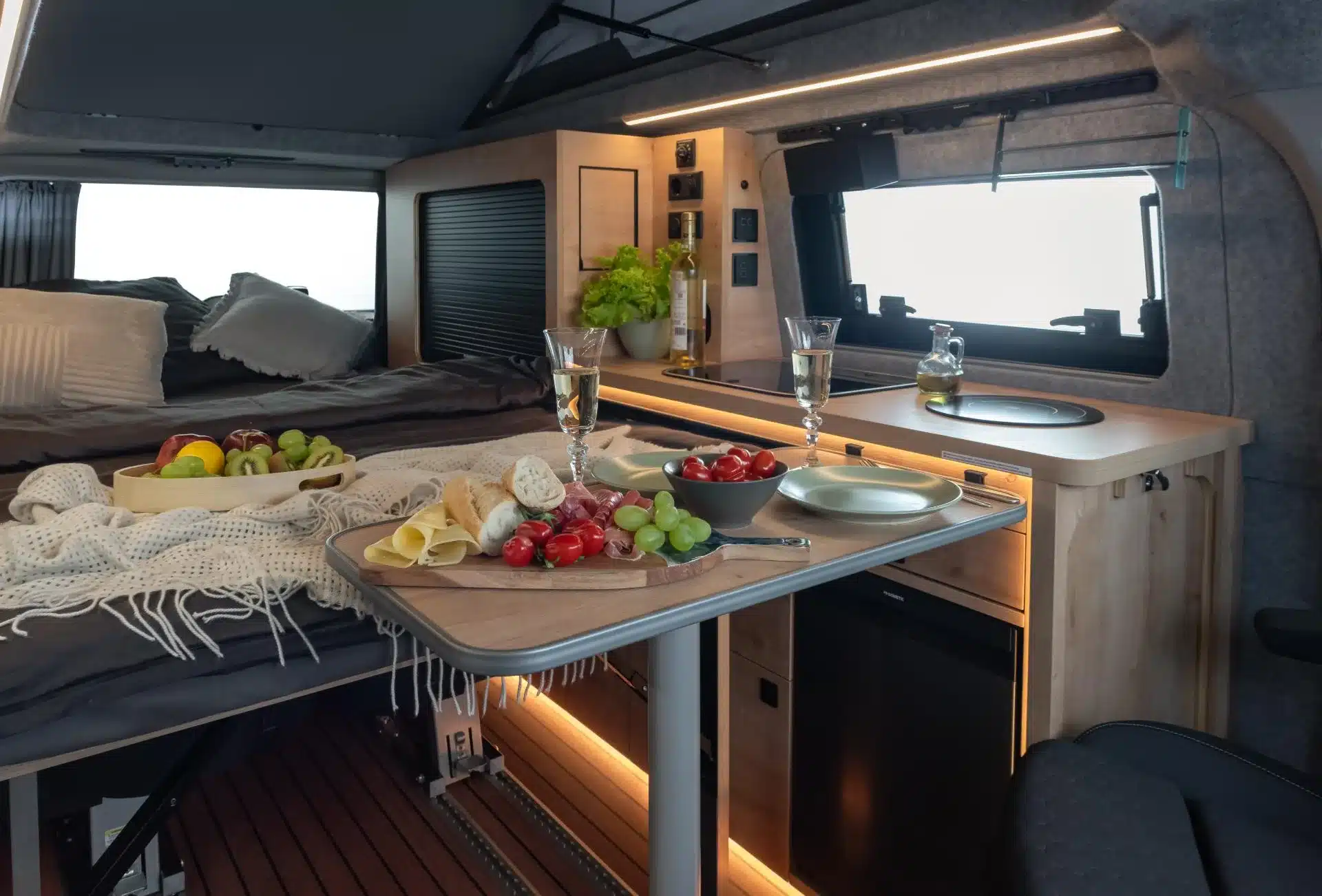 Innenraum eines Campervans mit Bett, Küchenzeile, Induktionskochfeld, Spülbecken und Tisch