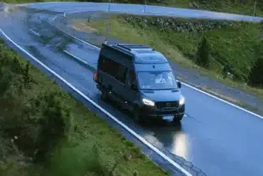 Dunkler Campervan auf einer nassen kurvenreichen Bergstraße vor einer alpinen Berglandschaft