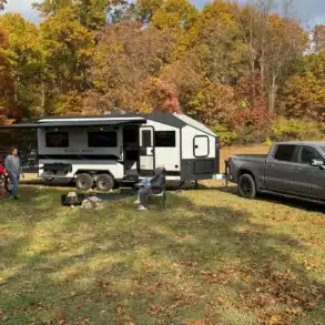 Black Wing 826-X Toy Hauler mit ausgefahrener Markise, Pickup und Offroad-Fahrzeugen auf einem herbstlichen Gelände