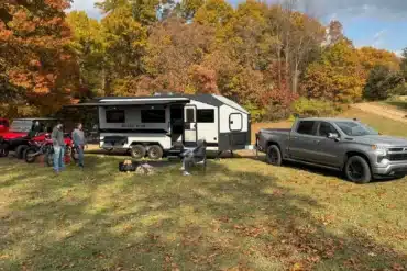 Black Wing 826-X Toy Hauler mit ausgefahrener Markise, Pickup und Offroad-Fahrzeugen auf einem herbstlichen Gelände