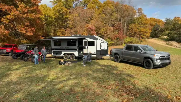 Black Wing 826-X Toy Hauler mit ausgefahrener Markise, Pickup und Offroad-Fahrzeugen auf einem herbstlichen Gelände