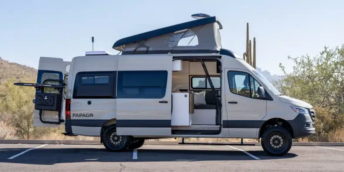 Weißer Papago Vans Campervan mit geöffnetem Aufstelldach, Heckträger und Schiebetür auf einem Parkplatz in einer Wüstenlandschaft.