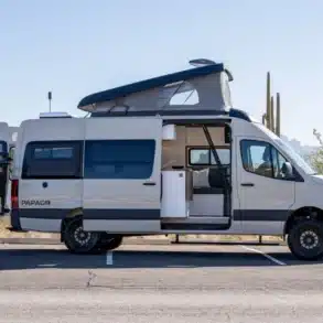 Weißer Papago Vans Campervan mit geöffnetem Aufstelldach, Heckträger und Schiebetür auf einem Parkplatz in einer Wüstenlandschaft.