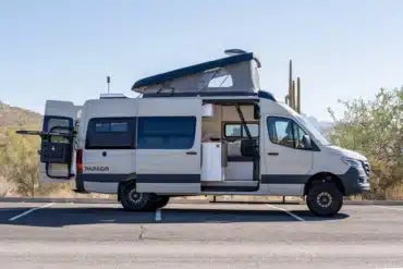 Weißer Papago Vans Campervan mit geöffnetem Aufstelldach, Heckträger und Schiebetür auf einem Parkplatz in einer Wüstenlandschaft.