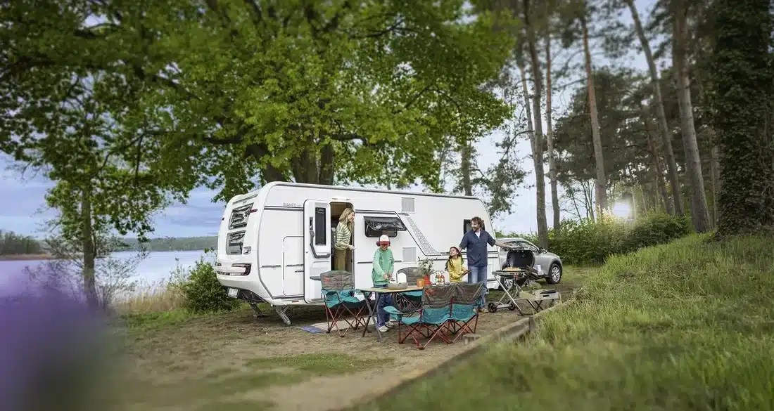 Weißer Fendt-Caravan BIANCO mit Familie und Campingmöbeln an einem See im Wald