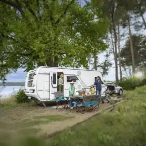 Weißer Fendt-Caravan BIANCO mit Familie und Campingmöbeln an einem See im Wald