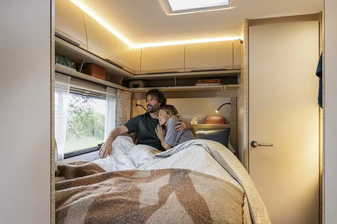 Innenraum eines Fendt-Caravan BIANCO mit Doppelbett, Küchen- und Stauraummöbeln sowie einem Paar im Schlafbereich