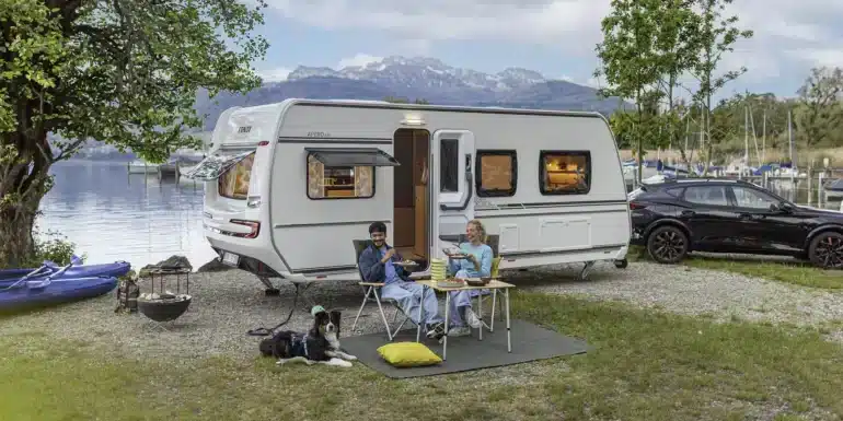 Weißer Fendt-Caravan APERO 515 mit geöffneter Tür am See vor einer Berglandschaft