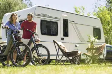 Weißer Fendt-Caravan TENDENZA 465 mit zwei Fahrrädern, Campingstühlen und Tisch auf einer Wiese.