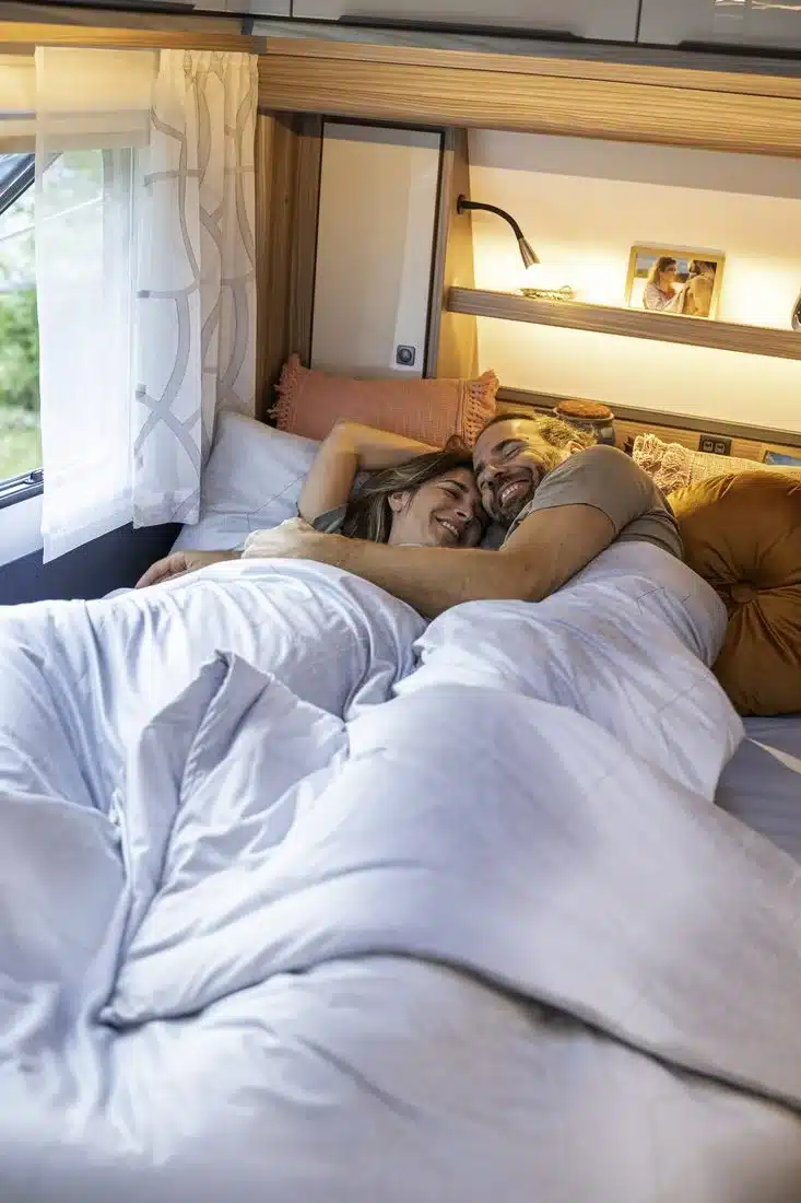 Paar liegt in einem hellen Doppelbett im Schlafbereich eines modernen Caravans