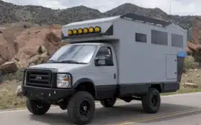 Grauer Field Adventure Vehicle Expeditionscamper mit Allradantrieb und Dachscheinwerfern auf einer Straße vor felsiger Landschaft