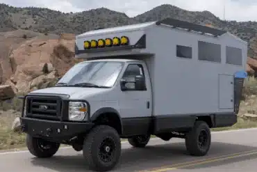 Grauer Field Adventure Vehicle Expeditionscamper mit Allradantrieb und Dachscheinwerfern auf einer Straße vor felsiger Landschaft