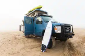 Blauer Field Van Ford Classic 4x4 mit Dachzelt, Surfbrett und Offroad-Stoßfänger auf Sand