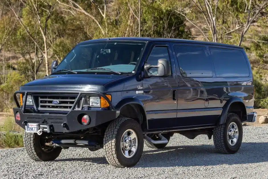 Blauer Ford E-350 Quigley 4x4 mit Offroad-Reifen und Frontstoßstange auf einem Schotterplatz