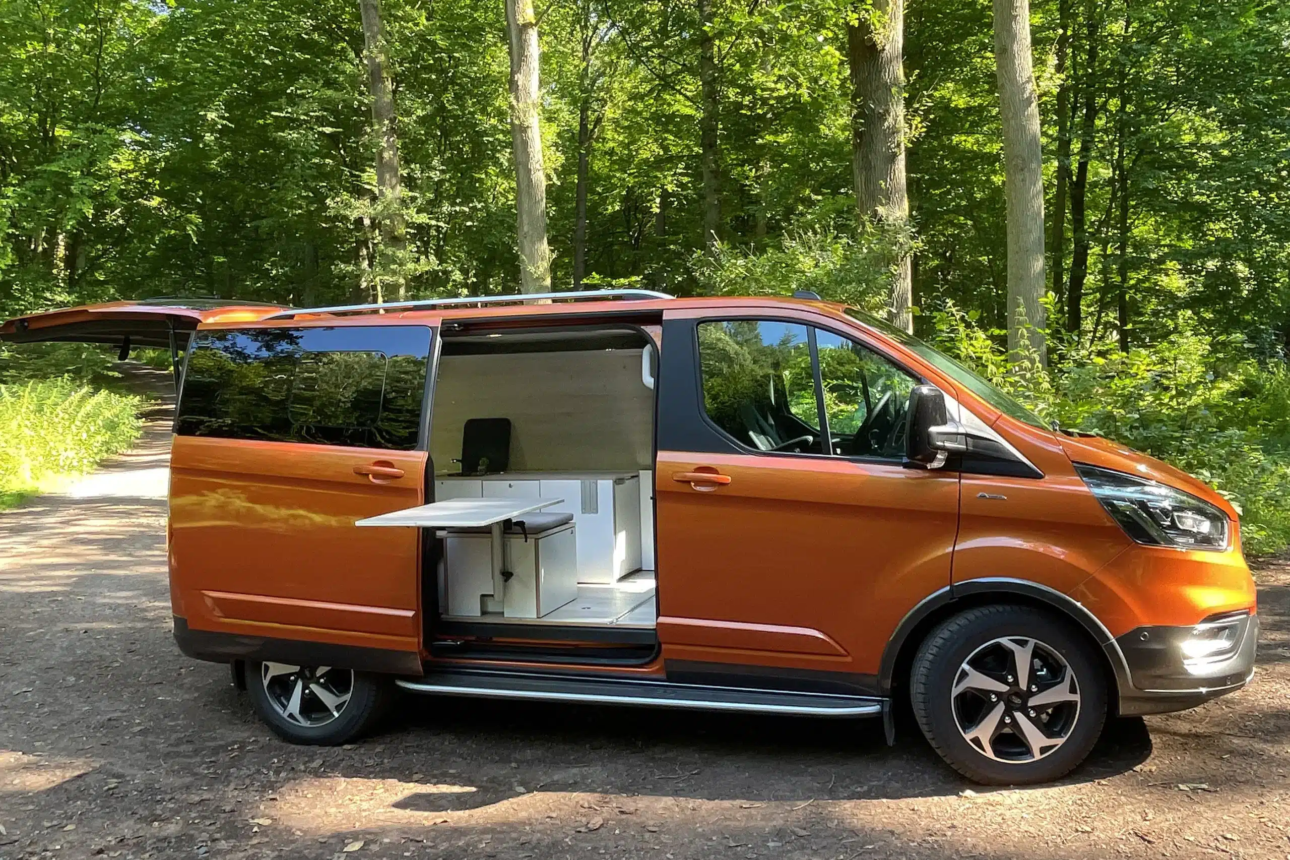 Orangefarbener Ford Transit Camper mit geöffnetem Seiteneinstieg, Küchenmodul und ausklappbarem Tisch