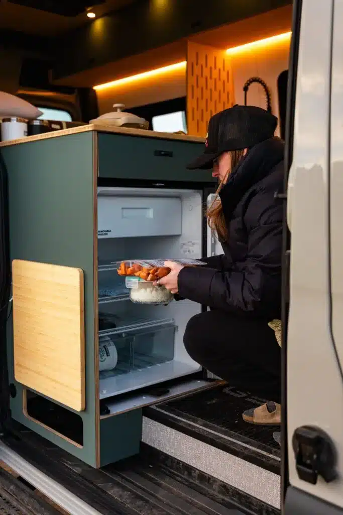 Person öffnet im Innenraum eines Campervans den Kühlschrank neben der Küchenzeile