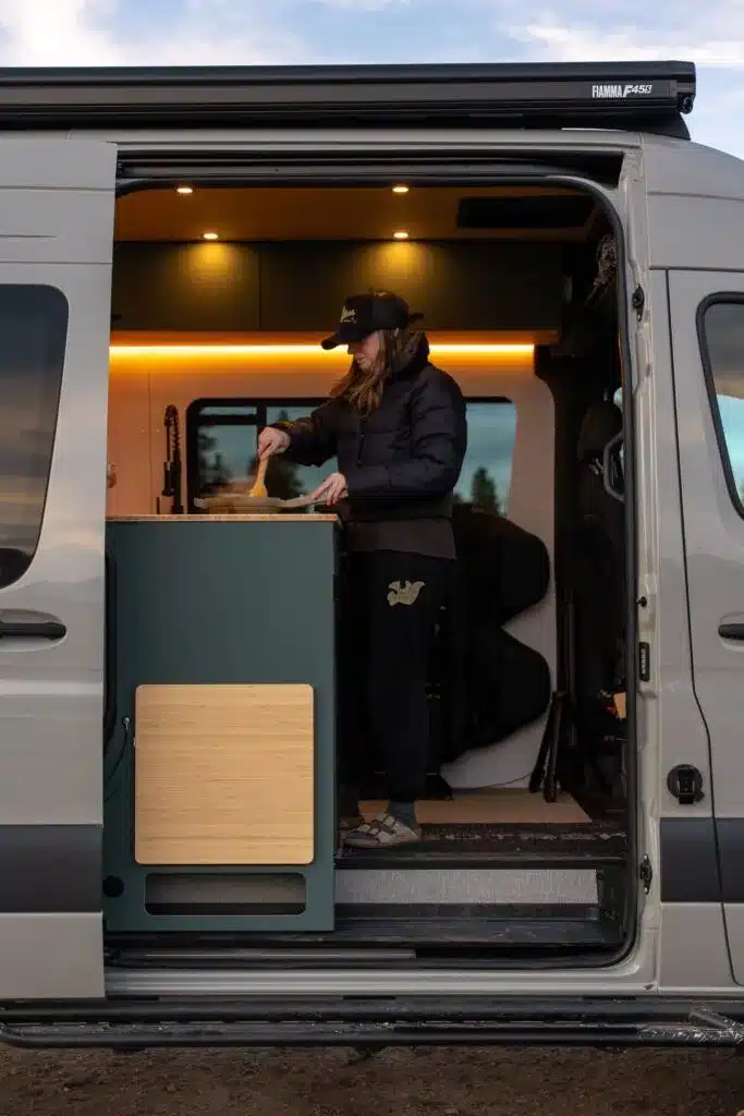 Innenraum eines grauen Geotrek Convoy Campervans mit Küchenzeile und Person beim Kochen
