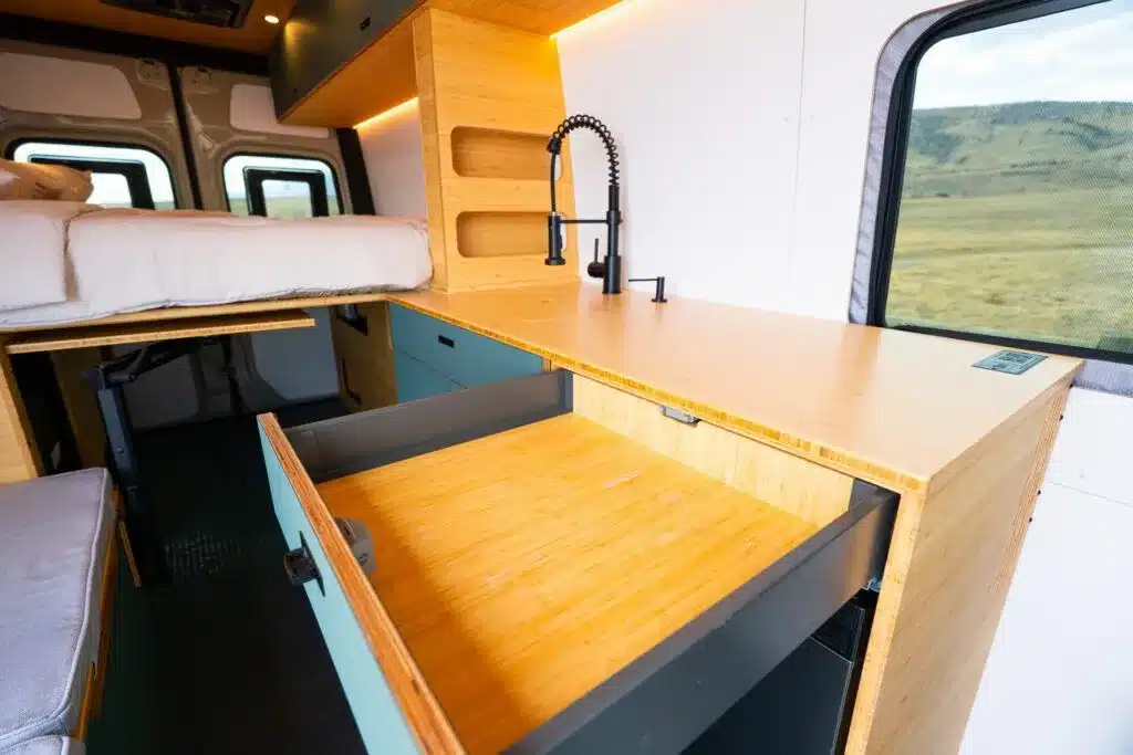 Innenraum des Geotrek Teton Campervans mit Küchenzeile, Spülbecken, Holzarbeitsfläche und längs ausgerichtetem Bett
