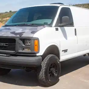 Weißer GMC Savana G2500 4x4 mit Höherlegung, großen Geländereifen und Camouflage-Motorhaube auf einer befestigten Straße.