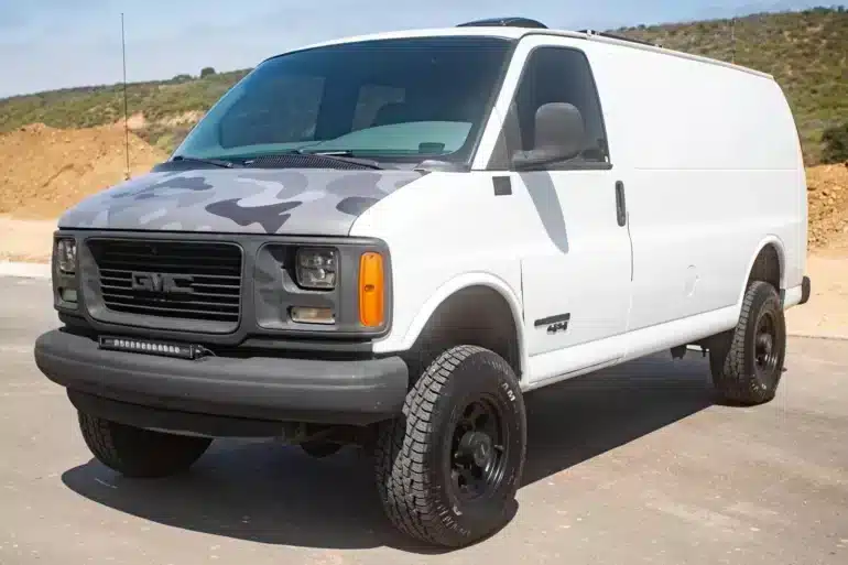 Weißer GMC Savana G2500 4x4 mit Höherlegung, großen Geländereifen und Camouflage-Motorhaube auf einer befestigten Straße.