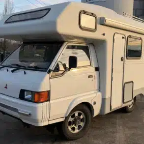 Weißer Mitsubishi Delica JB500 Camper in schräger Frontansicht mit Hochdach, Markise und Allradreifen