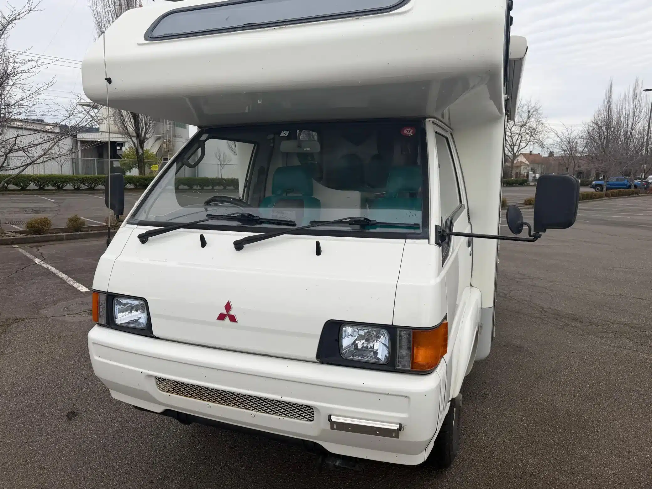 Weiße Frontansicht eines Mitsubishi Delica JB500 mit erhöhtem Camperaufbau auf einem Parkplatz