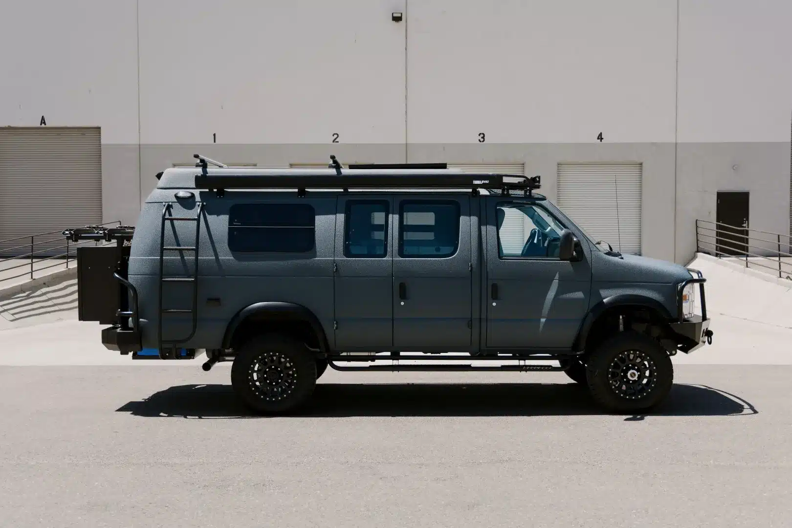 Dunkelgrauer Field Van Peak 4x4-Camper mit Dachträger, Leiter, Fahrradträger und groben Offroad-Reifen vor einem Gebäude.