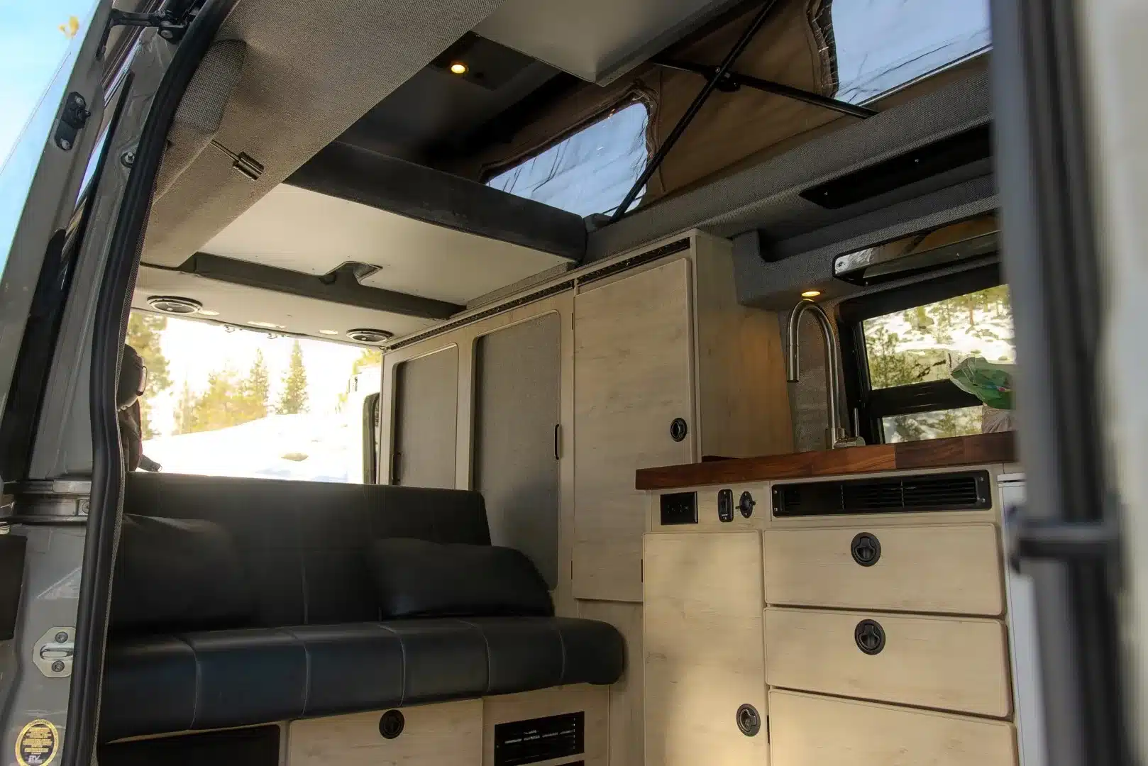 Innenraum eines Powers Sprinter Campervans mit geöffnetem Aufstelldach, schwarzer Sitzbank, Küchenzeile und Holzschränken