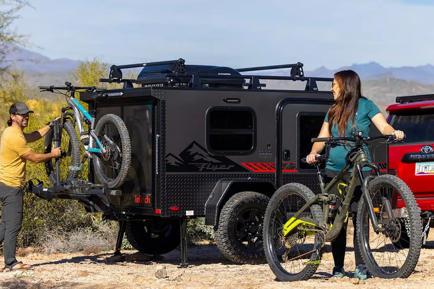 Schwarzer Flyer Pursue Offroad-Camper mit zwei Fahrrädern, Fahrradträger und rotem Zugfahrzeug in einer felsigen Landschaft
