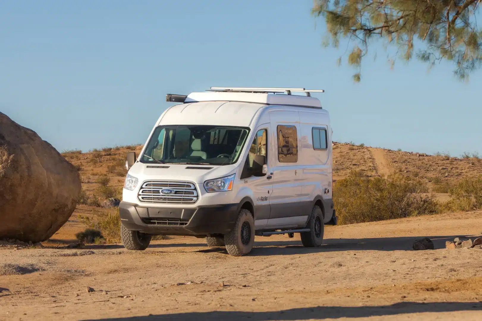 Weißer Tamarack-Camper auf Ford-Transit-Basis mit Pop-Top und Geländereifen in einer trockenen Landschaft