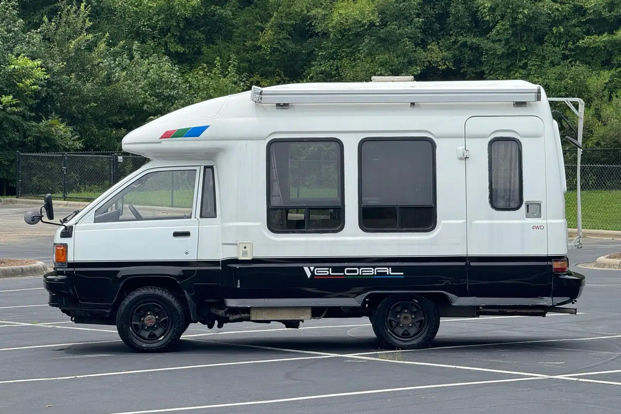 Weiß-schwarzer Toyota LiteAce Camper von 1990 mit Markise, Seitenfenstern und 4WD-Schriftzug auf einem Parkplatz