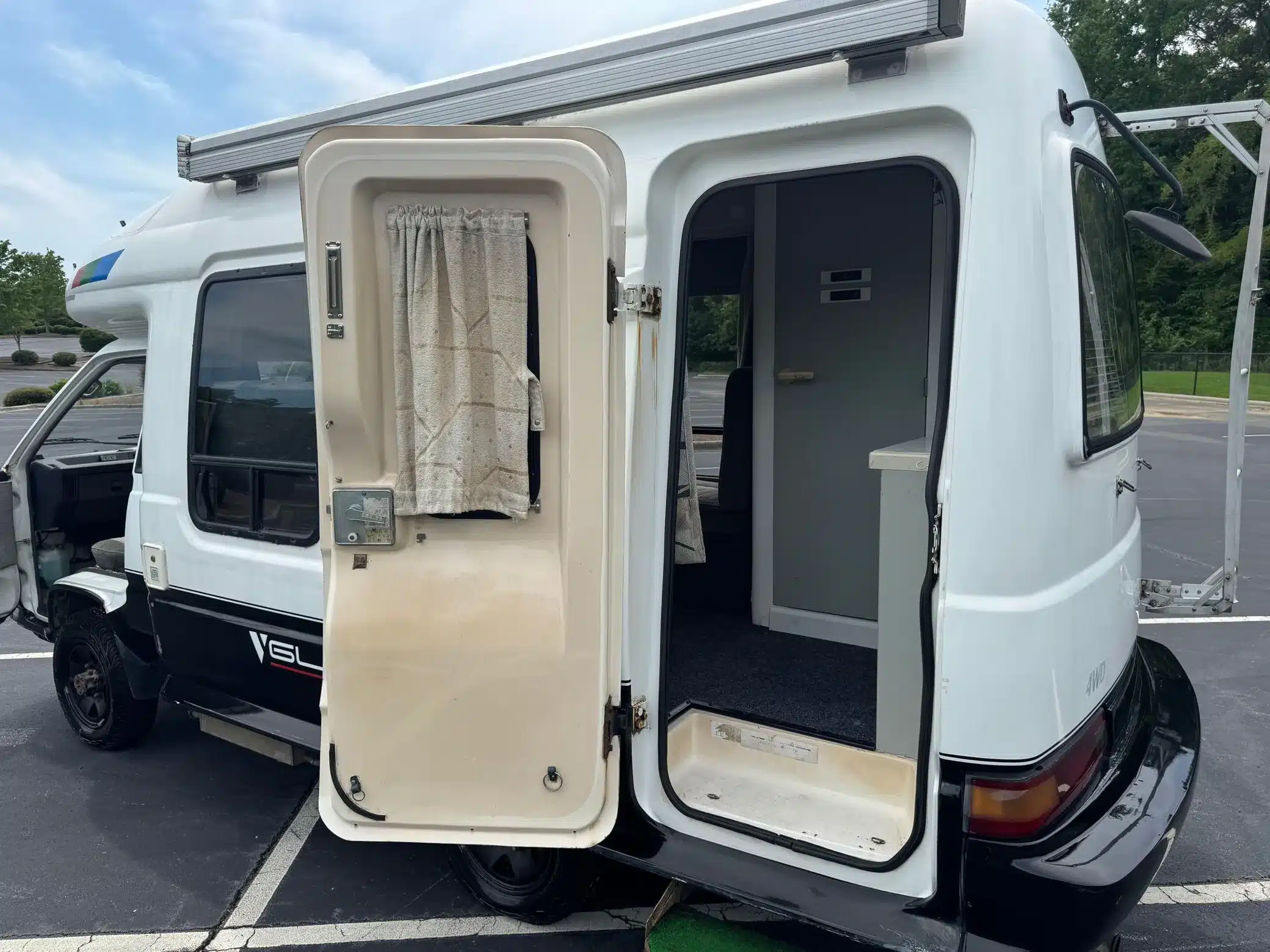 Weiß-schwarzer Toyota LiteAce Camper von 1990 mit geöffneter Seiten- und Hecktür auf einem Parkplatz