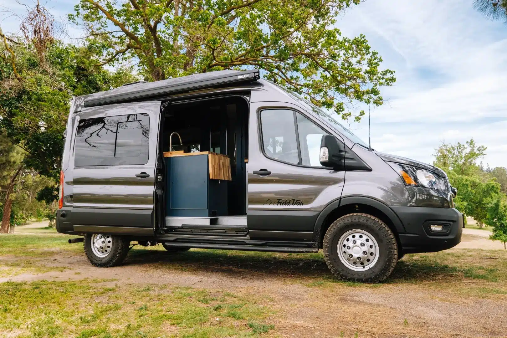 Grauer Field Van Tuolumne Campervan auf Ford-Transit-Basis mit geöffneter Schiebetür, Markise und grobstolligen Reifen